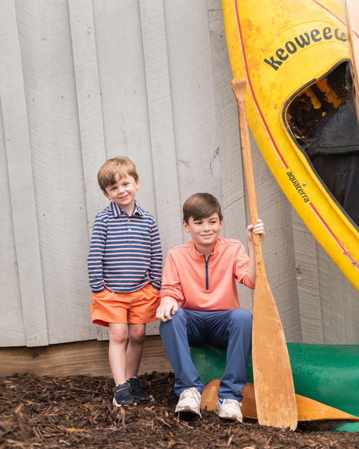 Two children standing next to a yellow canoe with an oar, against a wooden wall.