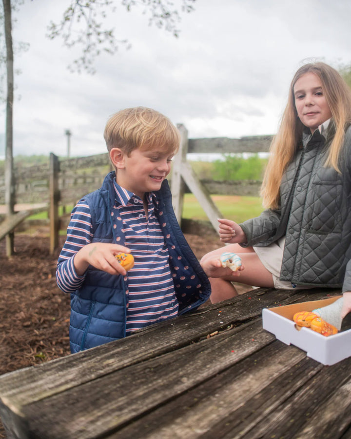 Two children at a wooden table outdoors, with one holding an orange and the other sitting.