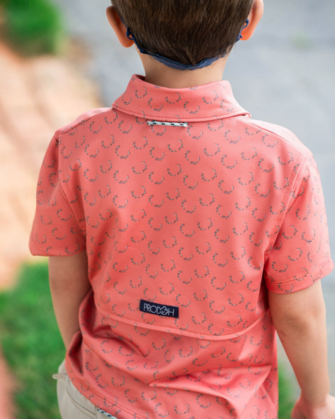 Child wearing a pink patterned shirt with a brand logo, standing outdoors.