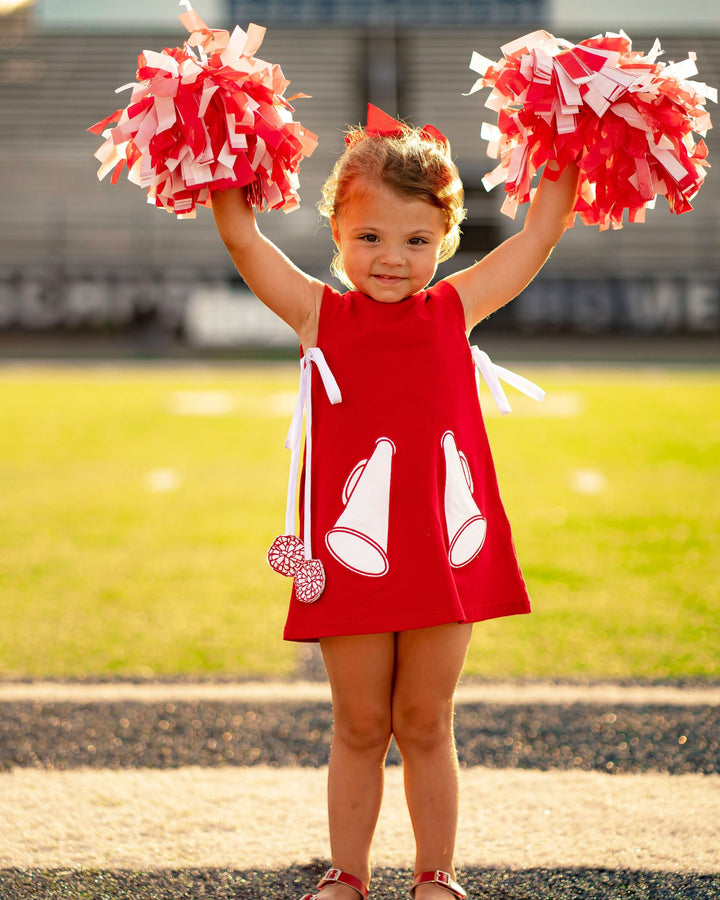 Child in a red dress with white megaphone designs, holding red and white pom-poms on a sports field.
