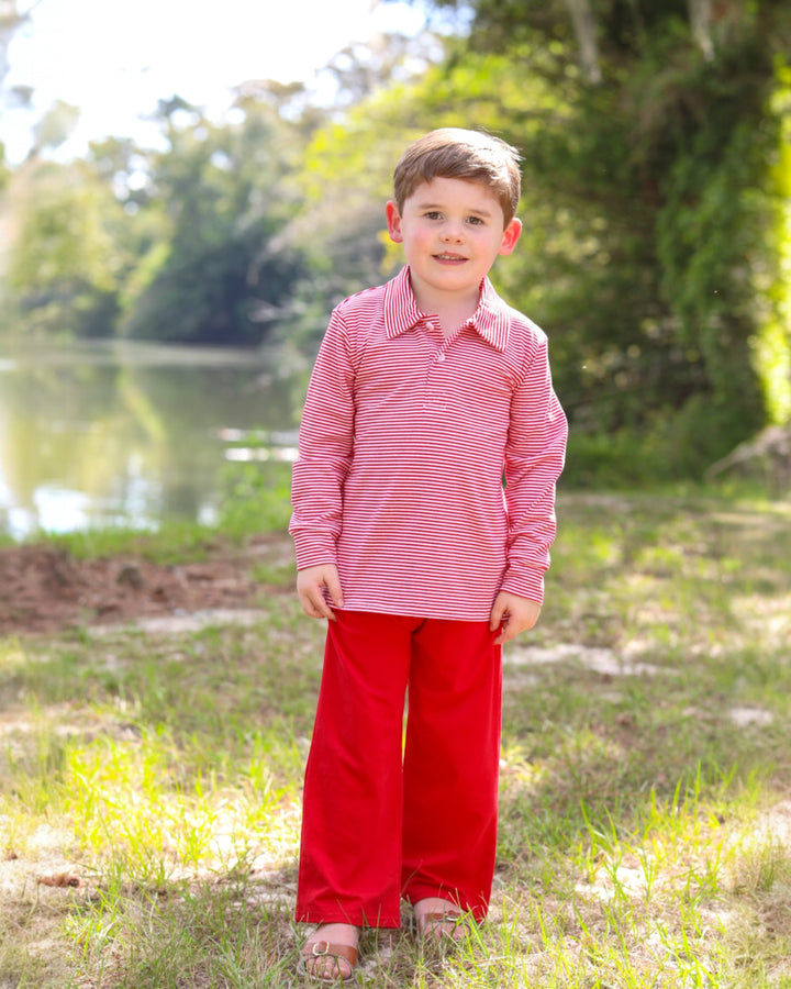 Young boy in a red and white striped shirt and red pants standing outdoors near a body of water.