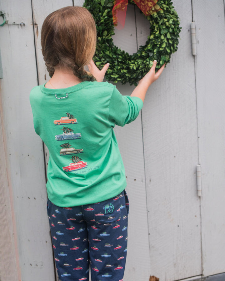 Child wearing a green shirt with vehicle designs and blue pants with car pattern, standing next to a green wreath on a wooden door.