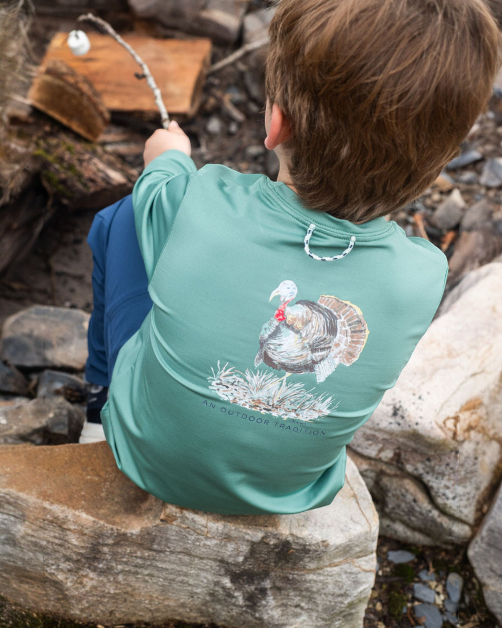Person wearing a teal shirt with a turkey design, sitting on rocks outdoors.