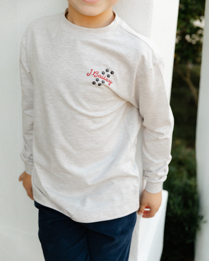 Child wearing a light gray long-sleeve shirt with a logo, standing against a white wall.