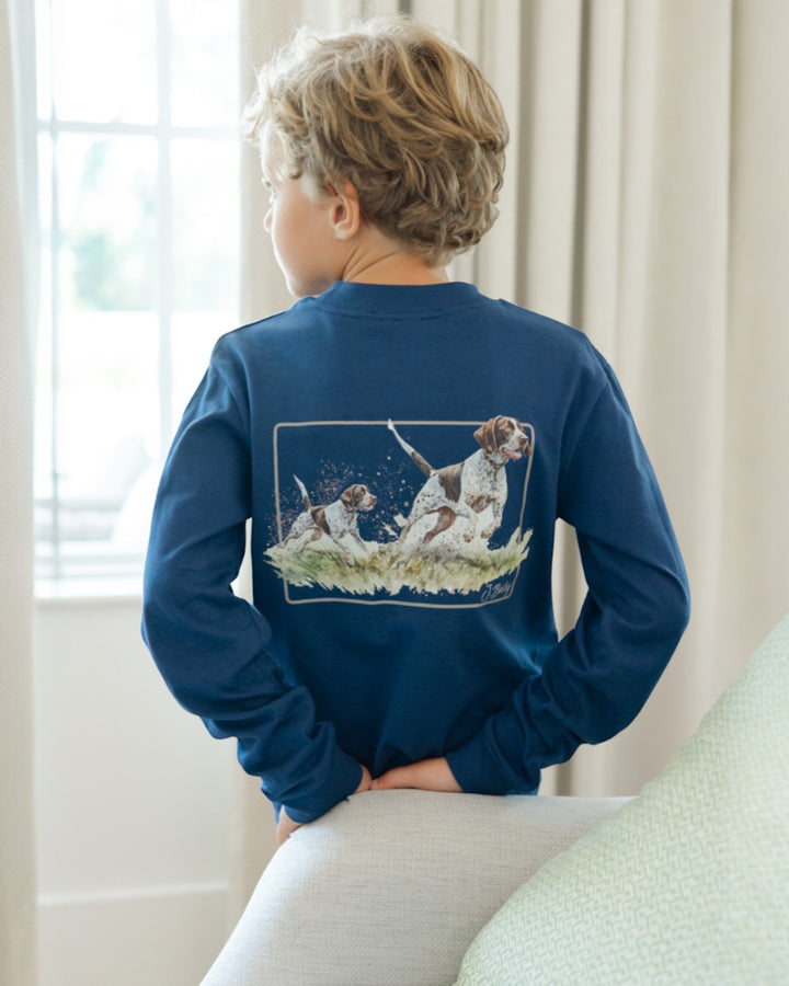 Child wearing a blue long-sleeve shirt with a graphic of dogs on the back, sitting in a room with light-colored curtains.