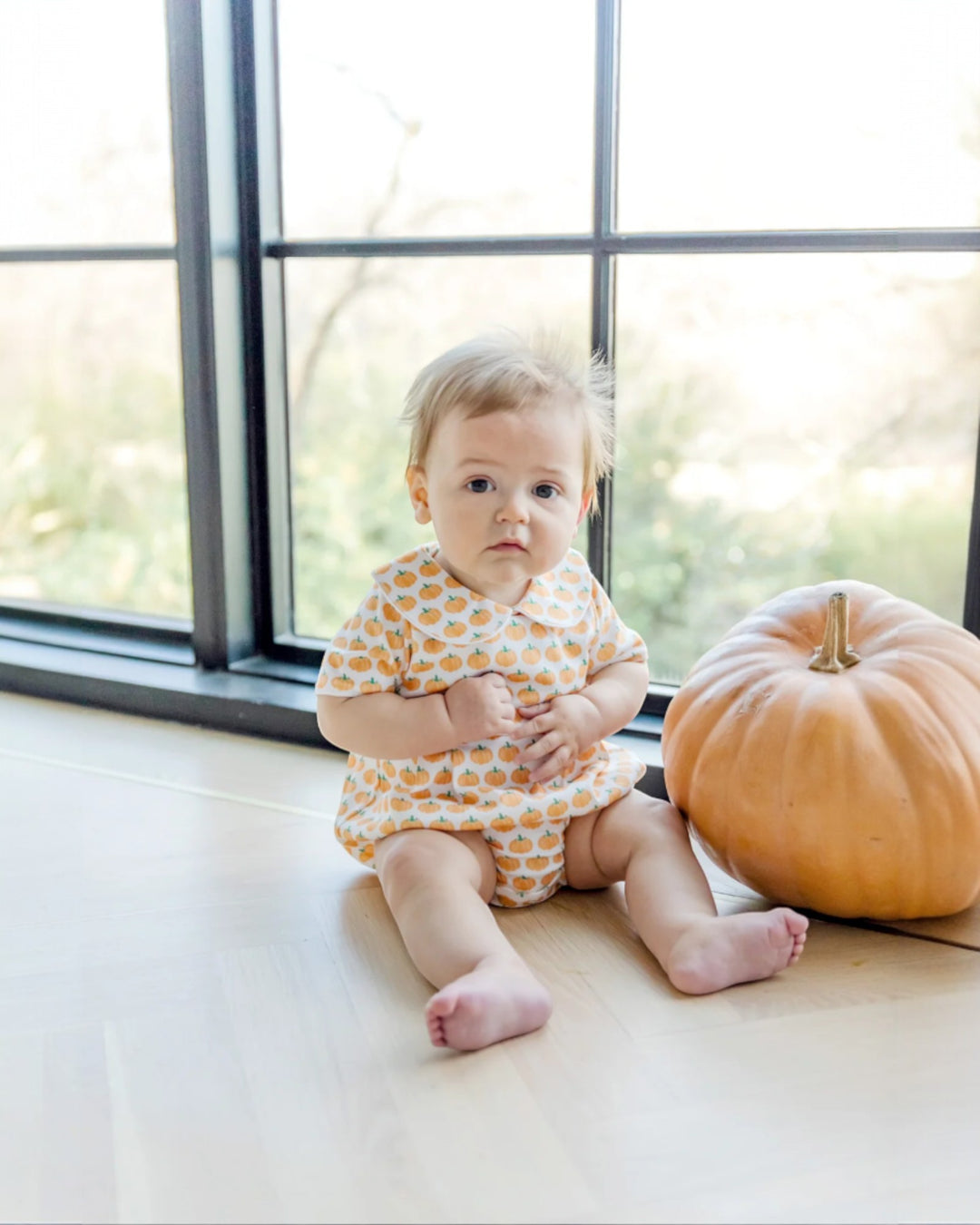 Baby in a polka dot outfit sitting next to a pumpkin in front of a window