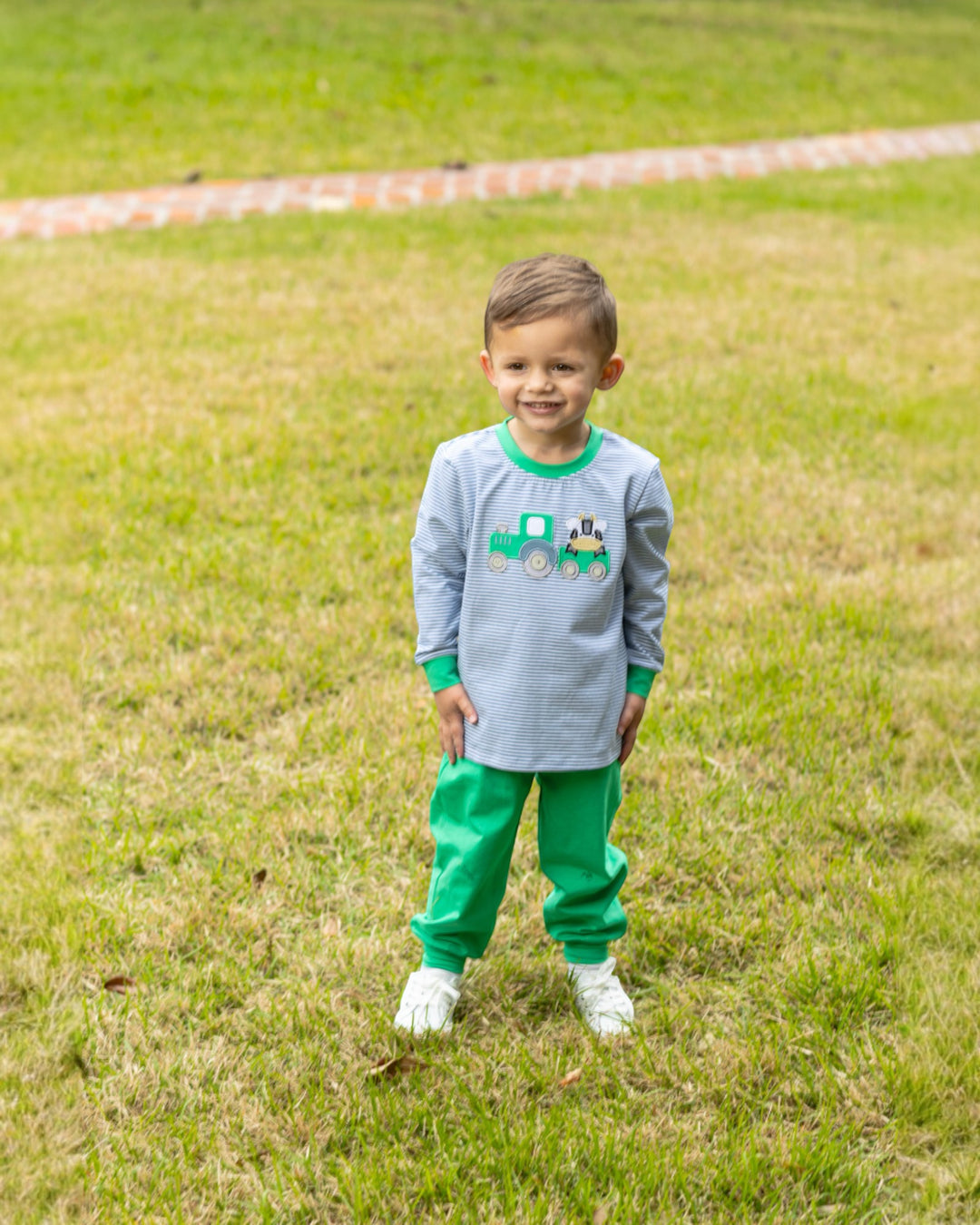 Child wearing a gray shirt with a green train design and green pants standing on grass.