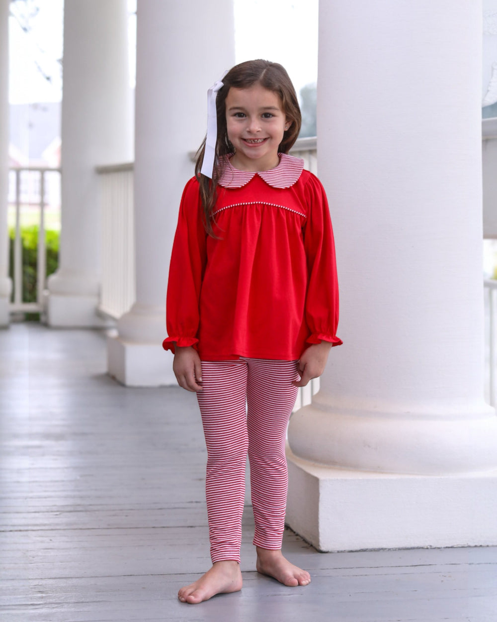 Young girl in a red top and pink striped leggings standing on a wooden floor with white columns in the background.