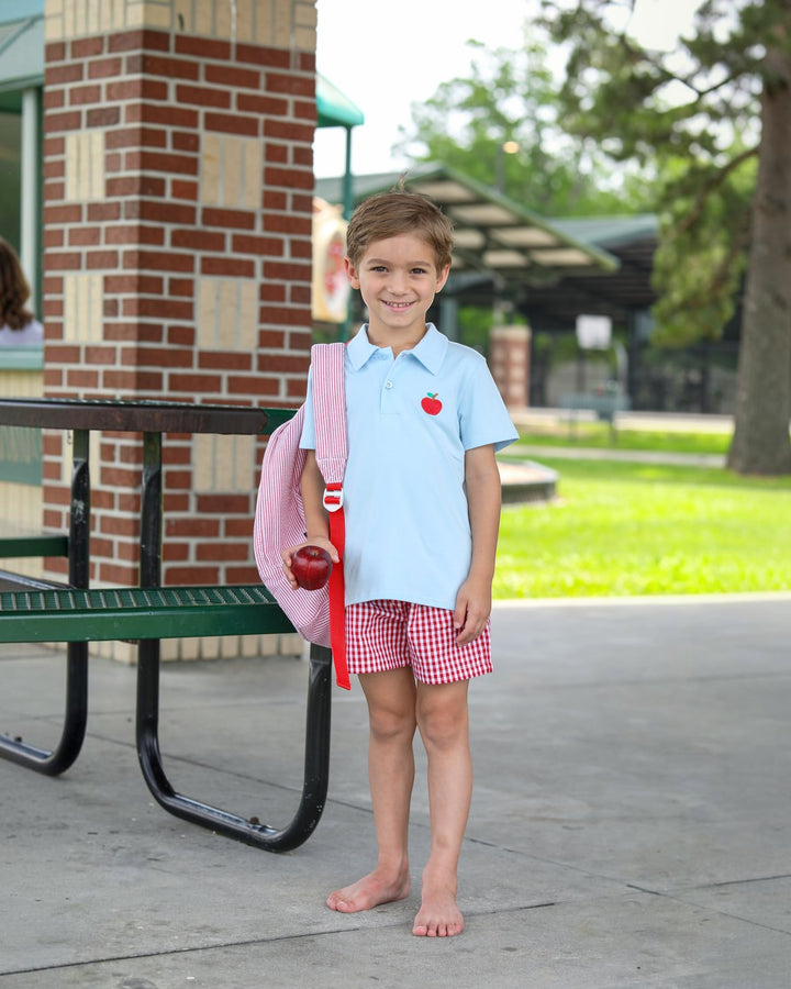 Child in light blue shirt and red checkered shorts holding a pink backpack and apple outdoors.