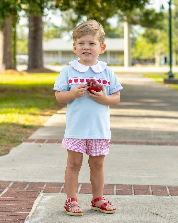 Child in light blue shirt with red trim and pink shorts holding an apple outdoors.