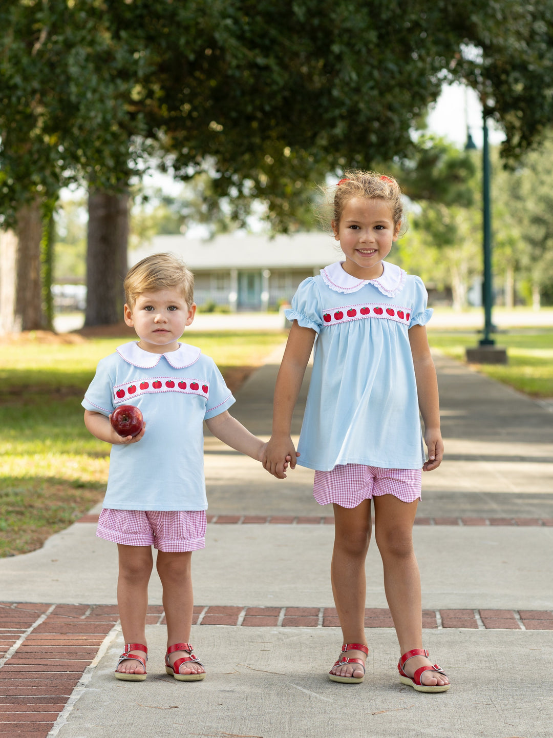 Two children in matching outfits standing on a sidewalk with trees in the background