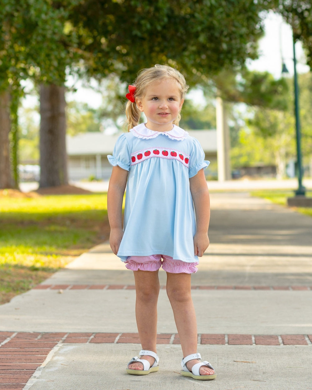 Young girl in a light blue dress with red trim standing on a sidewalk in a park.