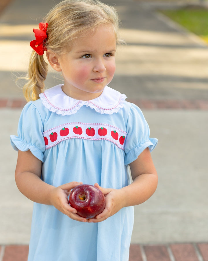 Young girl in a light blue dress with apple pattern holding an apple outdoors.