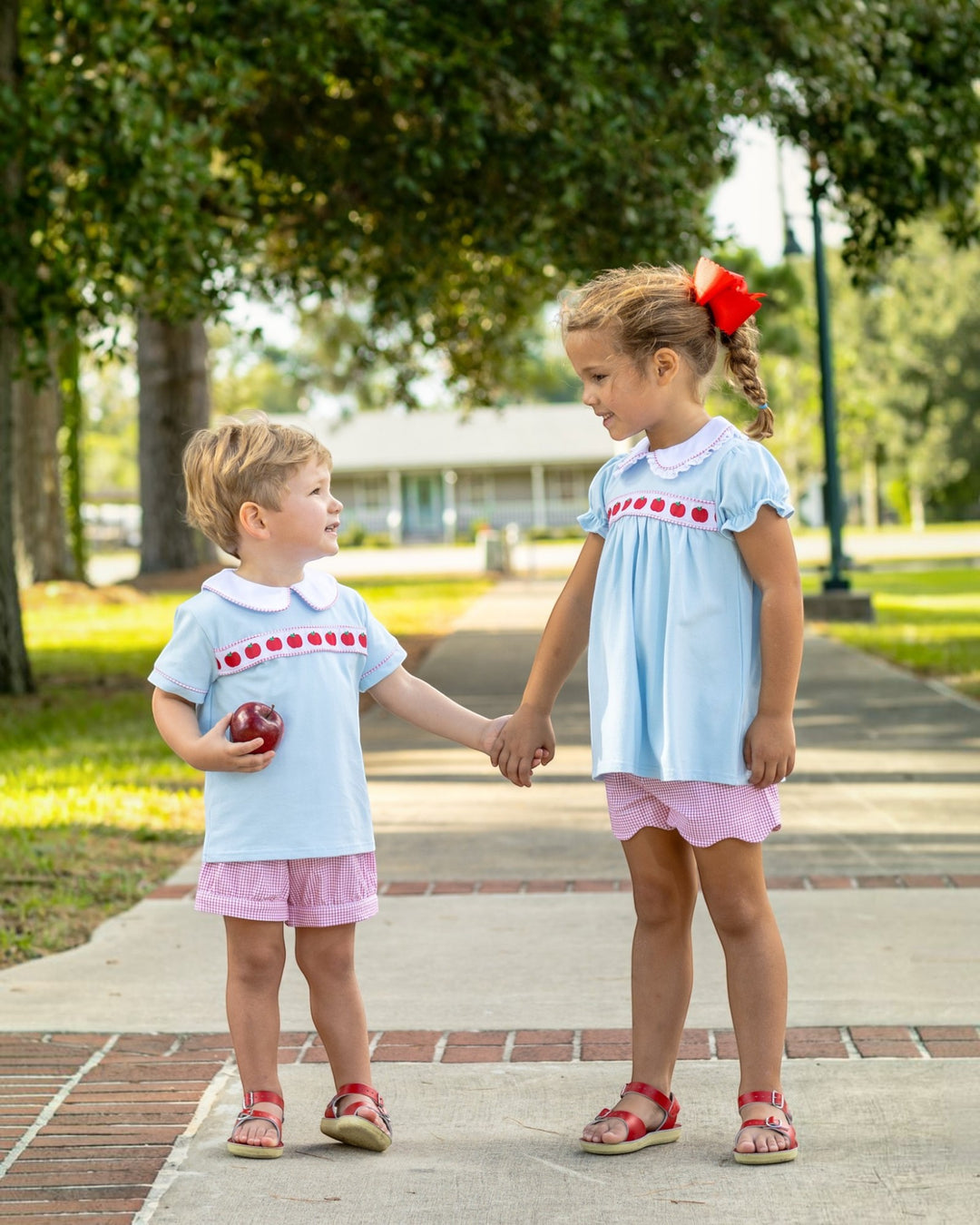 Two children holding hands on a sidewalk with trees and grass in the background