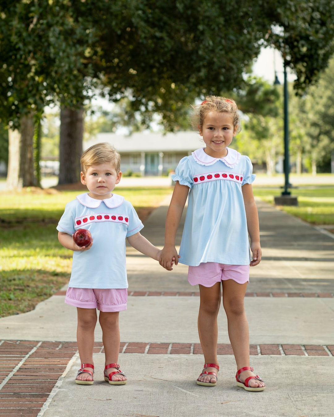 Two children holding hands on a sidewalk with trees and grass in the background