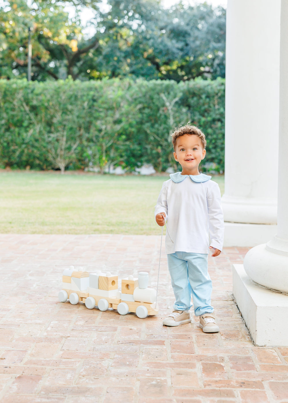 A young toddler stands on a brick porch next to a white column, holding a wooden toy train. The child is wearing a white long-sleeve top with a light blue collar, light blue pants, and beige sneakers, that store