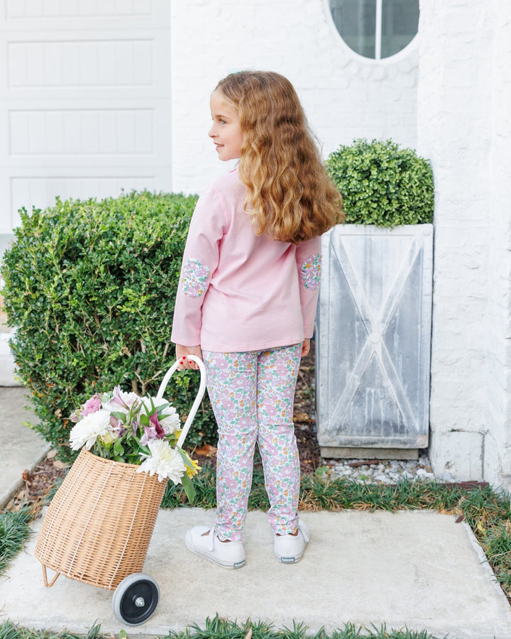 A young girl with long wavy light brown hair stands on a concrete walkway in front of a white house, wearing a pink long-sleeve top and matching floral print leggings. She is holding a wicker basket filled with white and pink flowers, that store