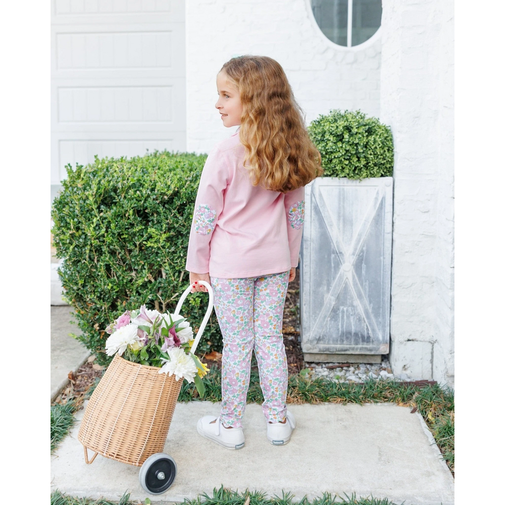 A young girl with long wavy light brown hair stands on a concrete walkway in front of a white house, wearing a pink long-sleeve top and matching floral print leggings. She is holding a wicker basket filled with white and pink flowers, that store