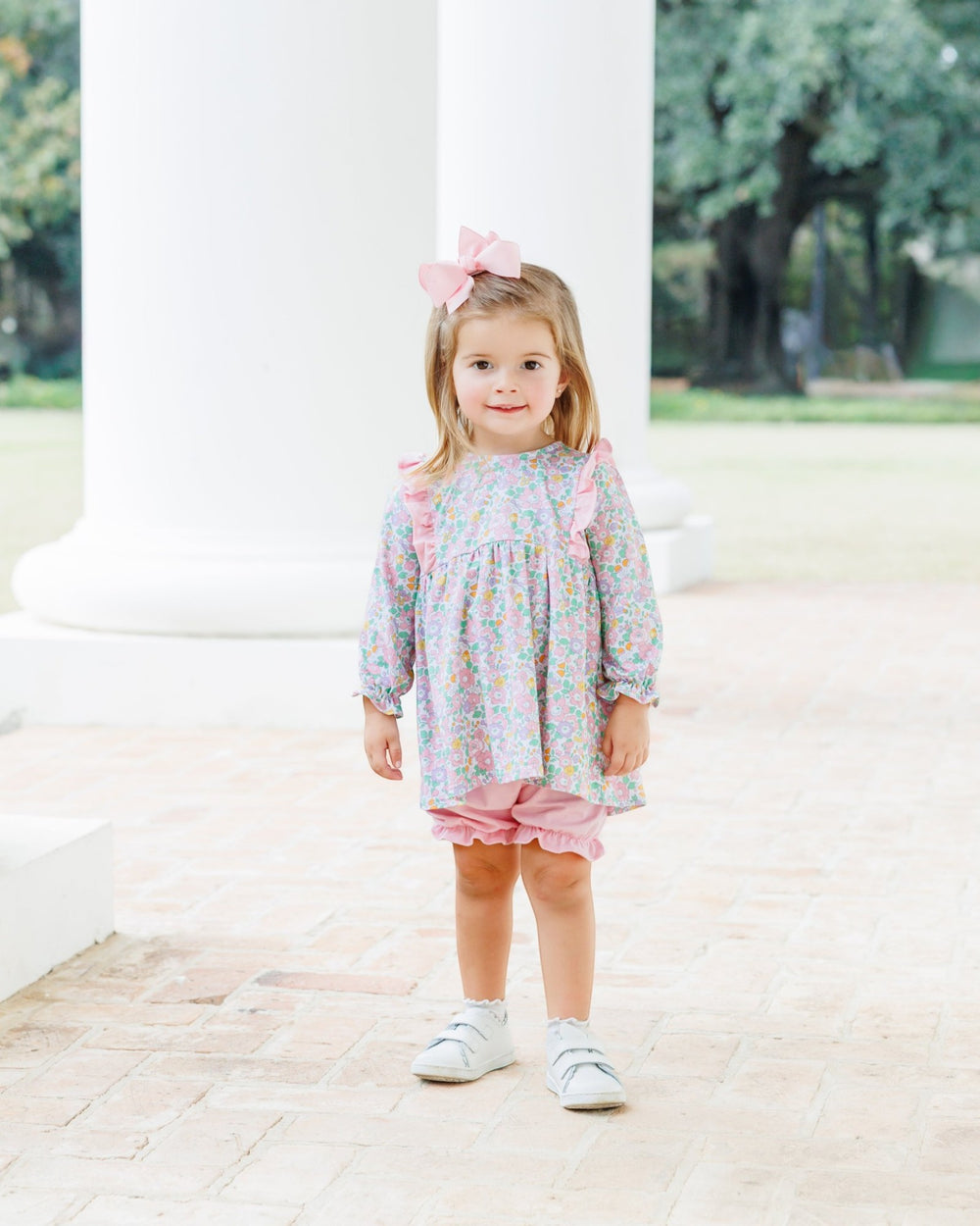 A young girl stands outdoors in front of white columns wearing a pastel floral long-sleeve top and pink ruffled shorts, with a pink bow in her hair and white sneakers, that store