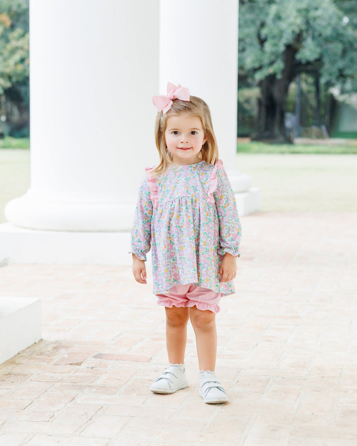 A young girl stands outdoors in front of white columns wearing a pastel floral long-sleeve top and pink ruffled shorts, with a pink bow in her hair and white sneakers, that store