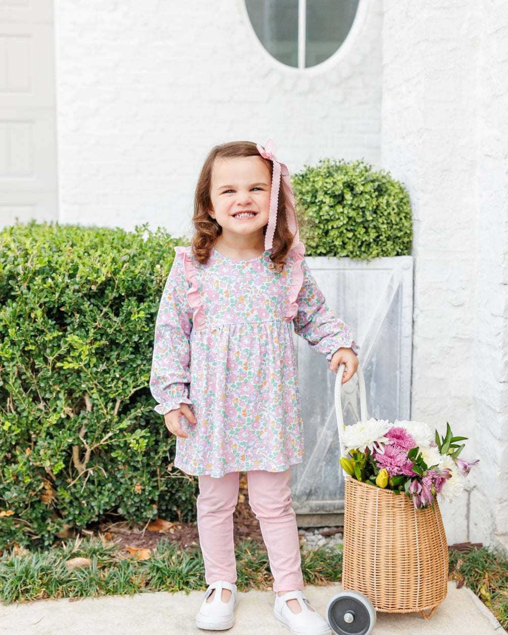 A young girl stands outdoors in front of a white house, wearing a pastel floral long-sleeve dress with pink ruffle trim and pink leggings, holding a wicker basket filled with flowers, that store