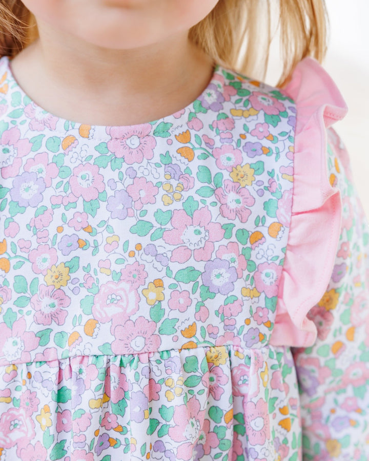 Close-up of a child wearing a long-sleeve dress with a pastel floral print in pink, green, yellow, and purple on a white background, featuring a ruffled neckline and ruffled sleeve, that store