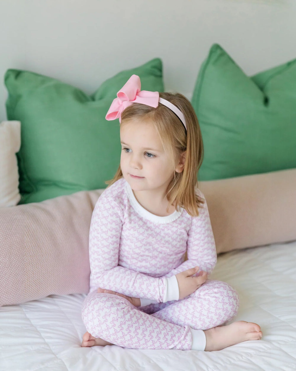 Young girl in pink pajamas with a bow sitting on a bed with green and beige pillows.