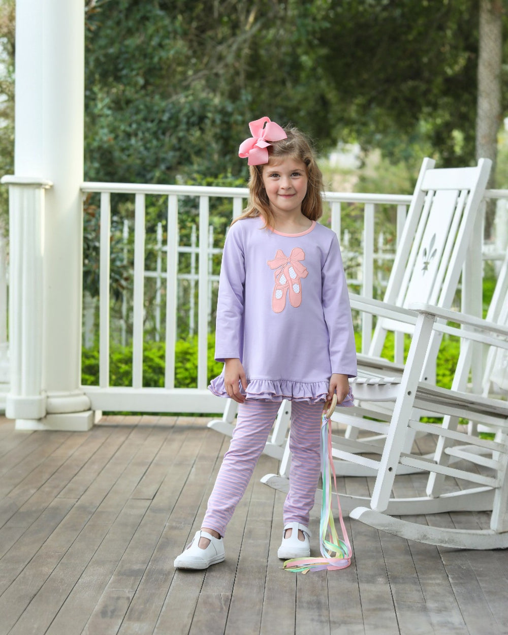 Young girl in a purple outfit standing on a wooden porch with white rocking chairs.