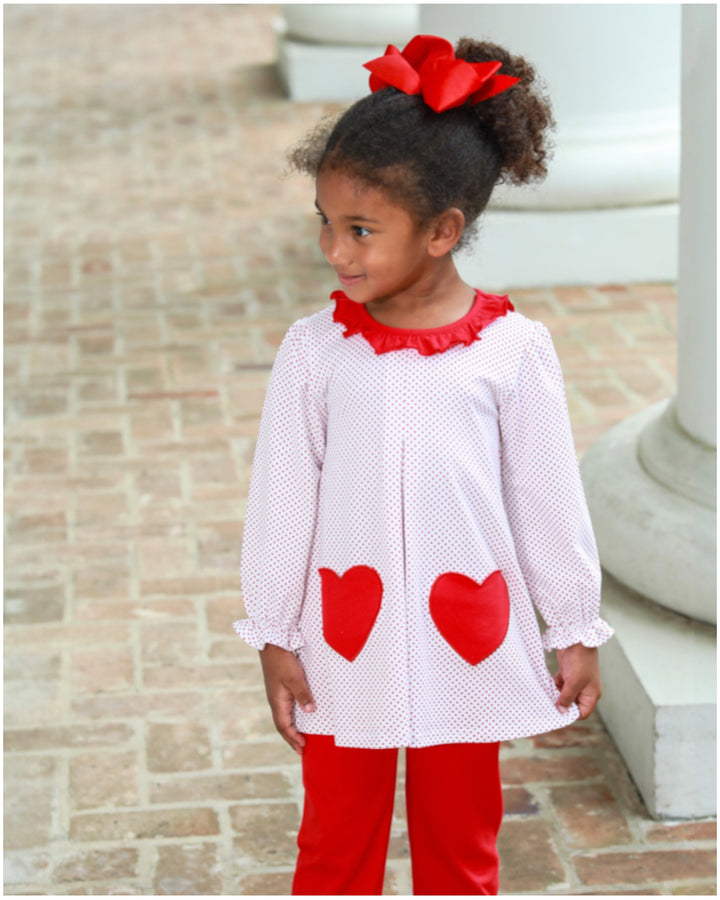 Young girl wearing a white dress with red hearts and red pants, standing on a brick surface.