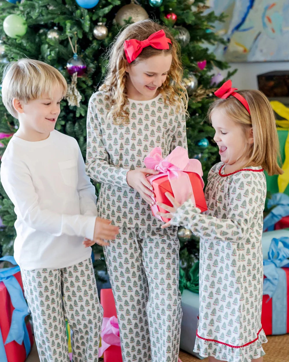 Three children in matching pajamas standing in front of a decorated Christmas tree.