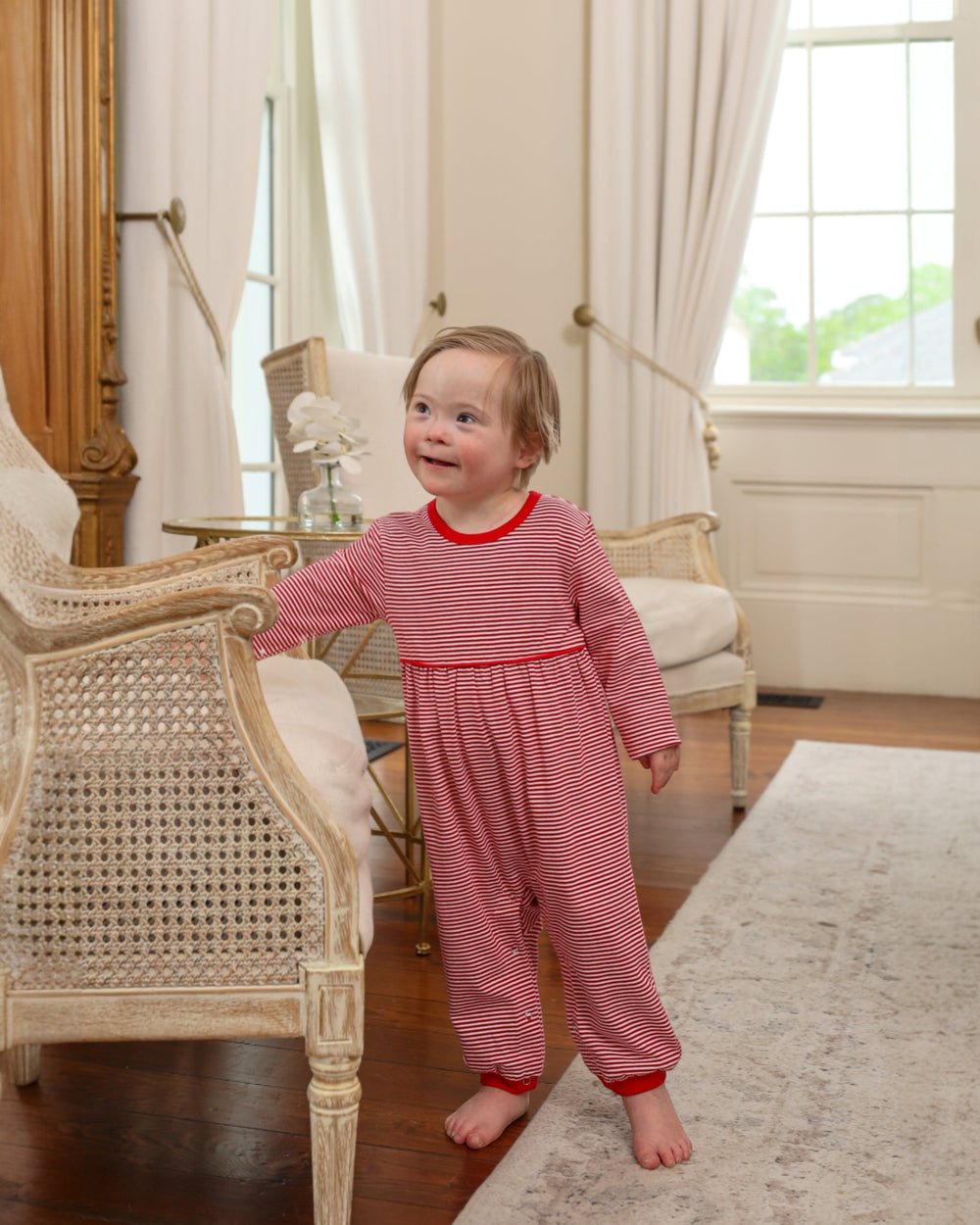 Child wearing a red and white striped outfit standing in a room with furniture and large windows.