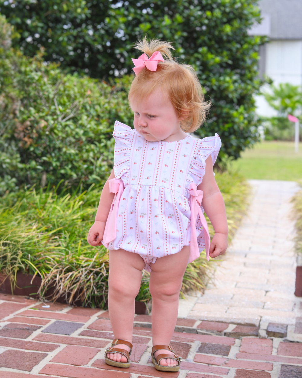 Young child in a pink and white outfit standing on a brick path with greenery in the background