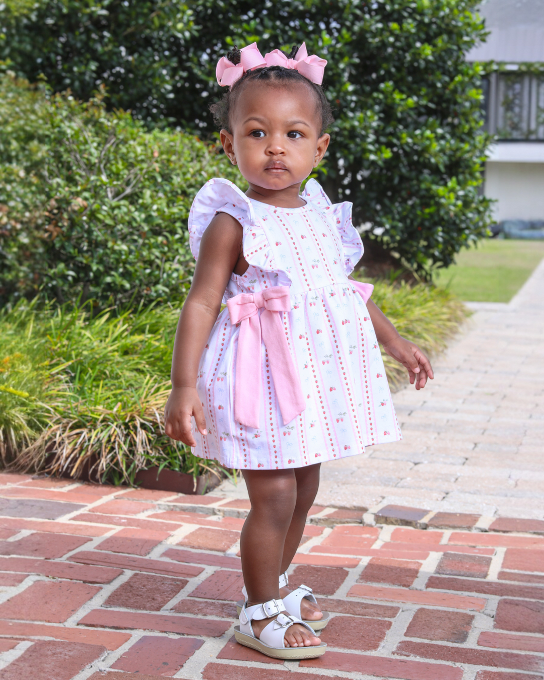 Young girl in a white dress with pink accents standing on a brick path.