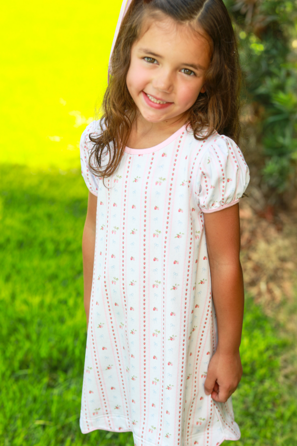 Young girl in a white dress with red embroidery standing in a grassy area.