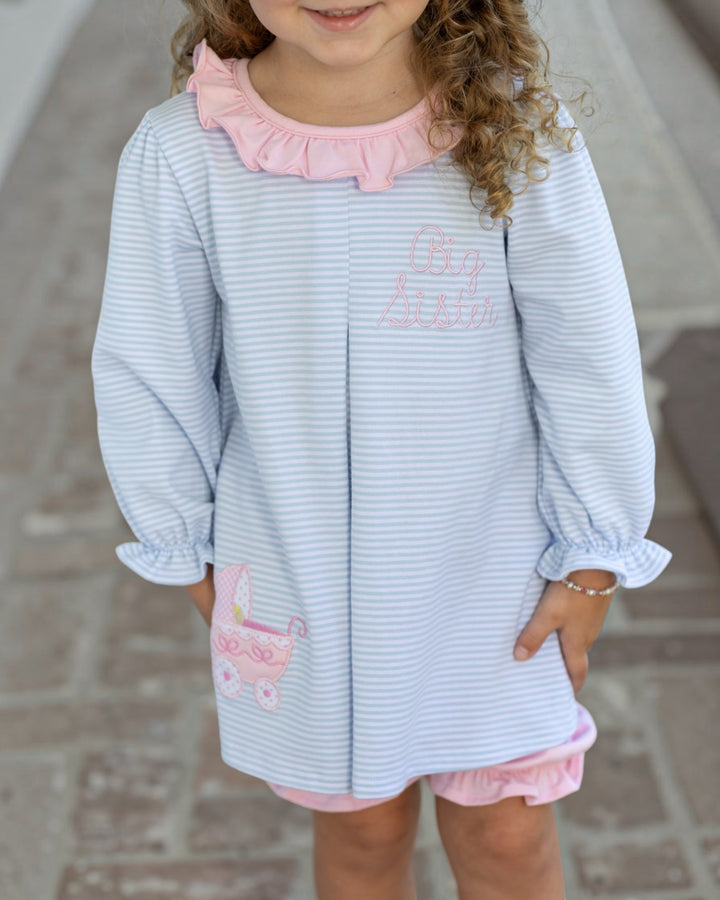 A young girl wearing a light blue and white striped long-sleeve tunic dress with pink ruffle trim, pink shorts, and pink embroidered details on the chest and pocket, standing outdoors on a paved walkway, that store