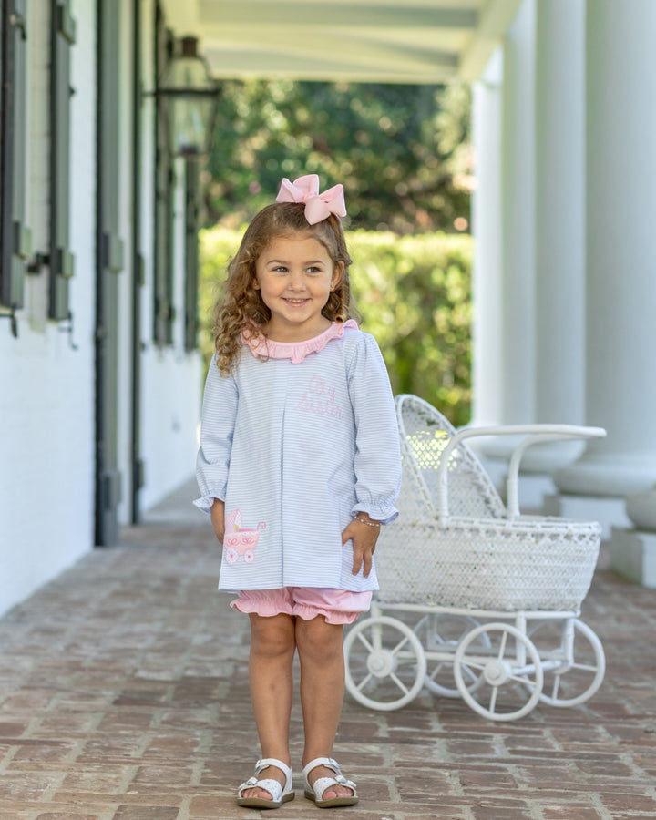 A young girl stands on a porch wearing a light blue and white striped long-sleeve tunic dress with pink ruffle trim, a pink bow in her hair, and pink shorts. A white wicker baby carriage is parked behind her, that store