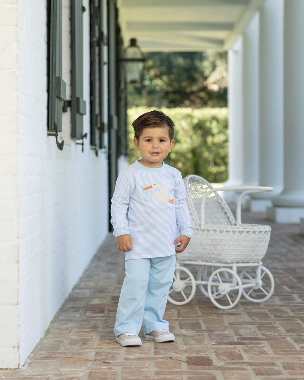A toddler stands on a porch wearing a light blue long-sleeve shirt with a duck graphic and light blue pants. A white wicker bassinet is beside him, with a white brick wall and columns in the background, that store