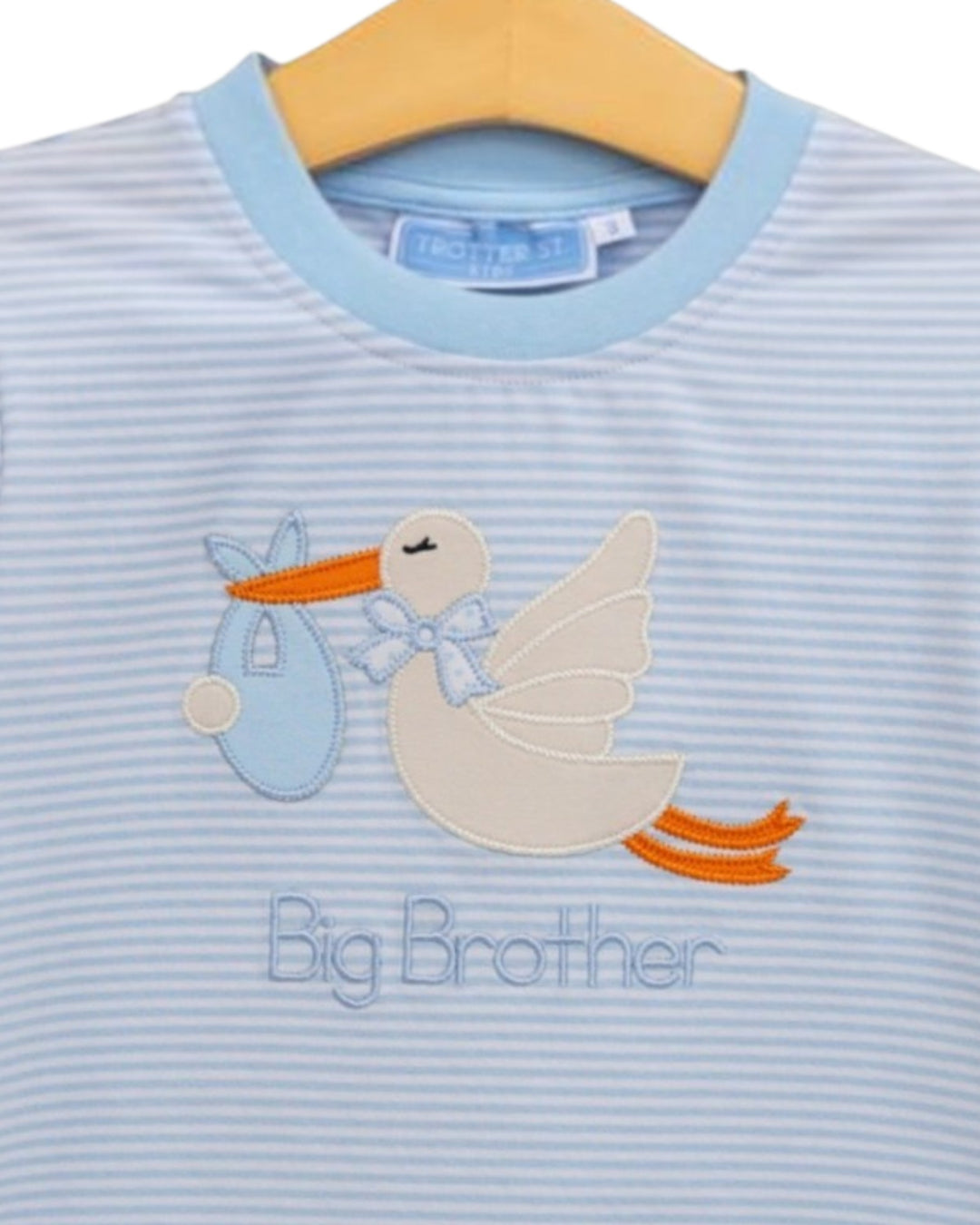 A close-up of a baby t-shirt with light blue and white horizontal stripes, featuring an embroidered stork carrying a blue baby bottle and the words &