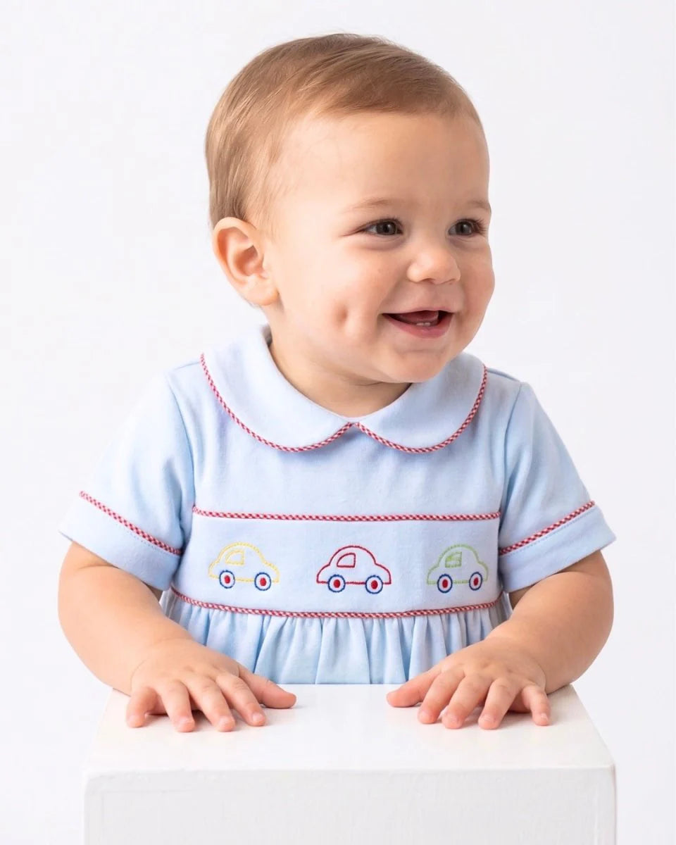 A baby wearing a light blue short-sleeve dress with a Peter Pan collar and embroidered car details, smiling and looking to the side while standing behind a white block, that store