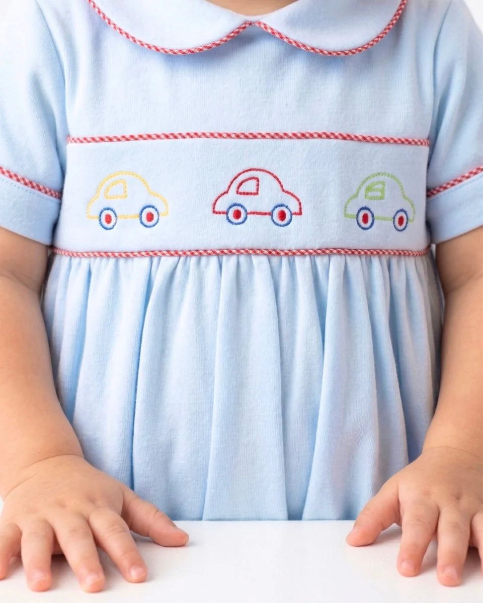 A close-up of a baby wearing a light blue short-sleeve dress with red stitched trim and embroidered cars across the chest, that store