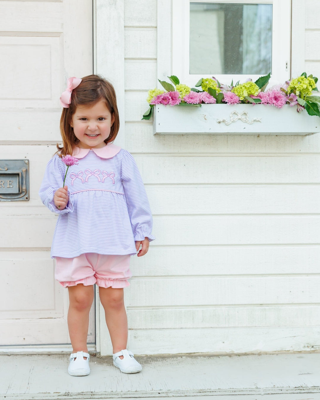 A young girl stands in front of a white house wall wearing a light lavender and pink striped long-sleeve top with a pink collar and embroidered detail, pink shorts, and white shoes. She has a pink bow in her hair and holds a small pink flower, that store