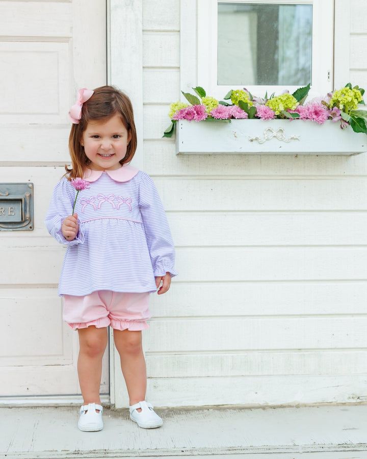 A young girl stands in front of a white house wall wearing a light lavender and pink striped long-sleeve top with a pink collar and embroidered detail, pink shorts, and white shoes. She has a pink bow in her hair and holds a small pink flower, that store