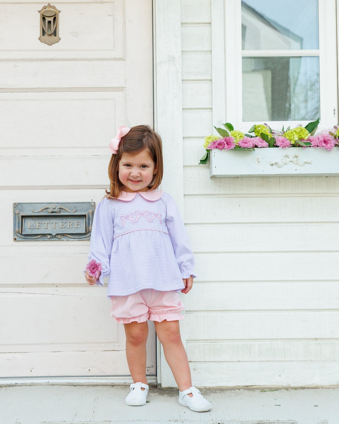 A young girl with a pink bow in her hair stands in front of a white house door wearing a light purple and pink striped long-sleeve top with a Peter Pan collar, pink shorts, and white shoes, that store