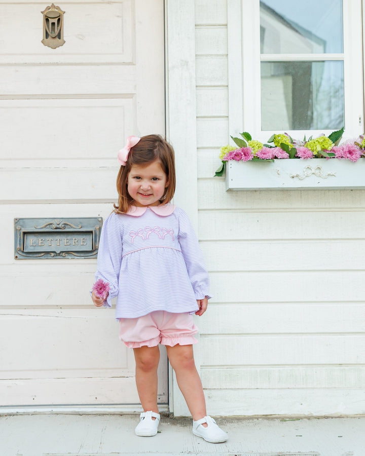 A young girl with a pink bow in her hair stands in front of a white house door wearing a light purple and pink striped long-sleeve top with a Peter Pan collar, pink shorts, and white shoes, that store