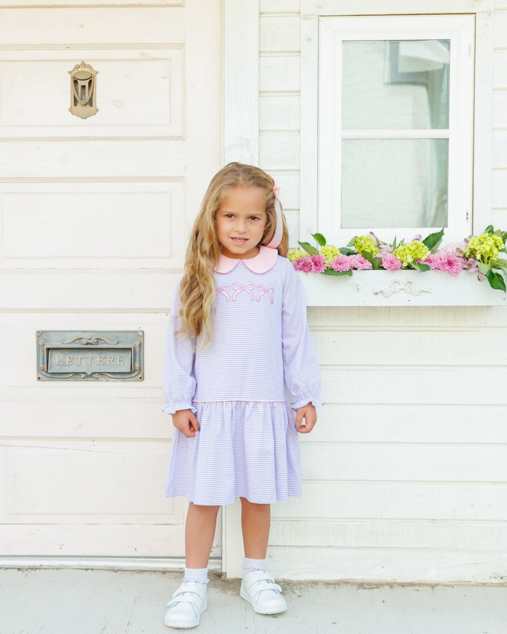 A young girl with long blonde hair stands in front of a white house wall wearing a light lavender and white gingham dress with a pink collar and embroidered chest detail, paired with white socks and white sneakers, that store