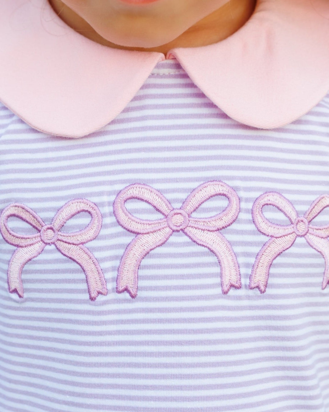 Close-up of a child wearing a light pink collar and a lavender and white striped top with three pink embroidered bows across the chest, that store