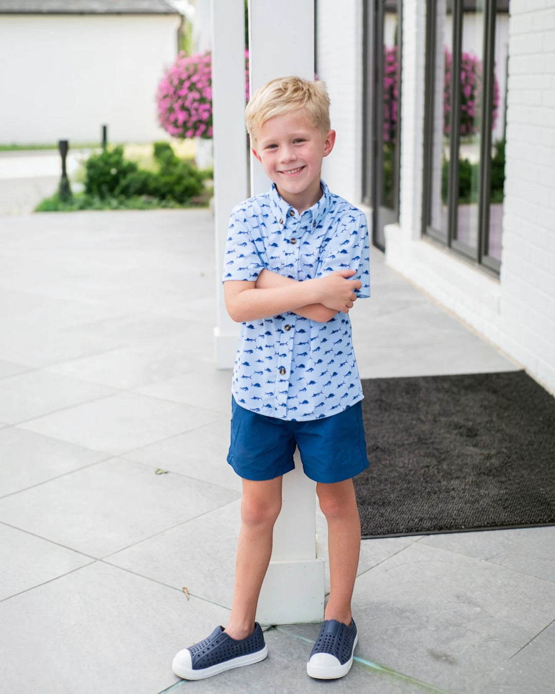 A young blond boy stands outdoors in front of a white building, wearing a light blue short-sleeve button-up shirt with a small repeating dark pattern, navy shorts, and navy slip-on shoes, that store