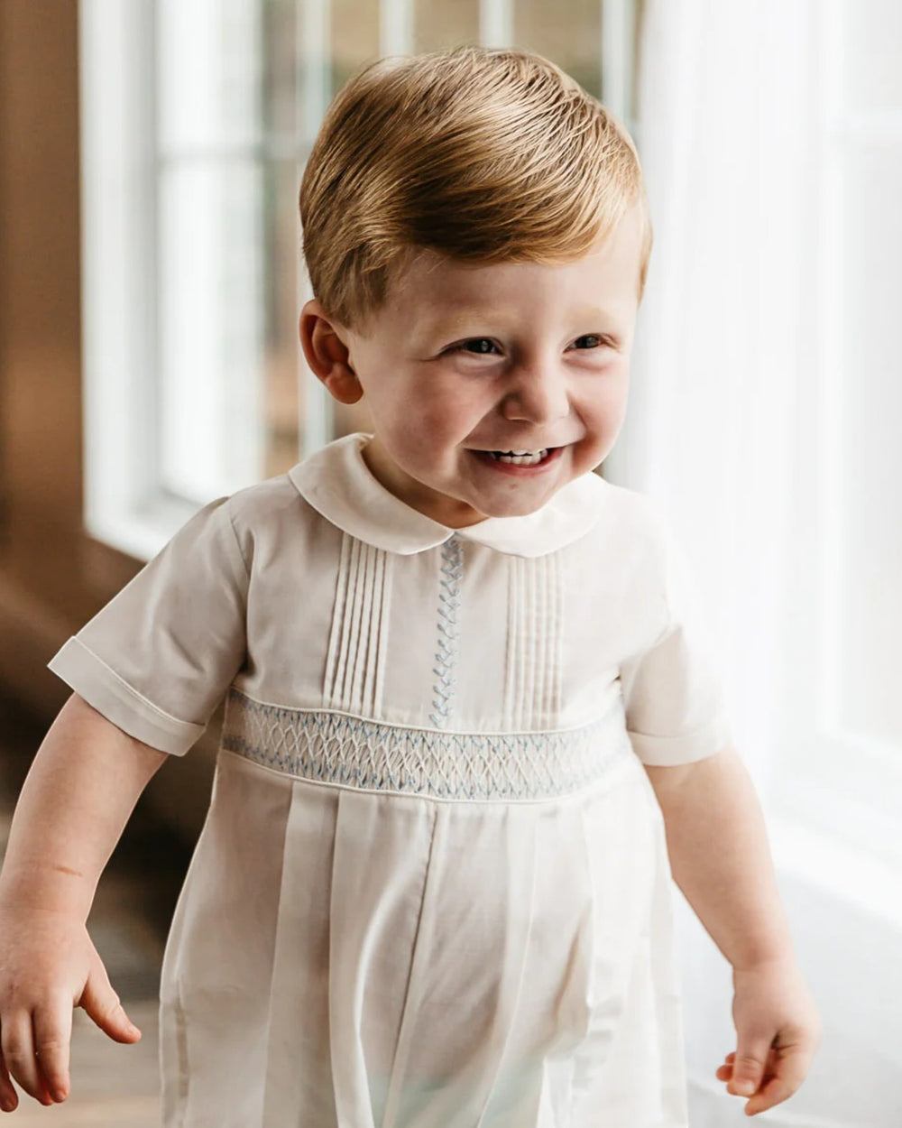 Child wearing a white dress with a lace collar in a softly blurred indoor setting