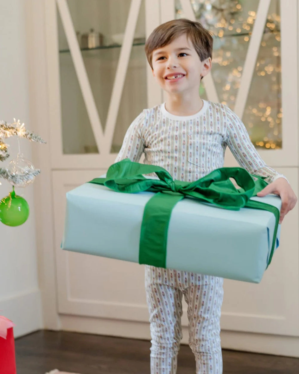 Child holding a large gift box with a green ribbon in a festive indoor setting.