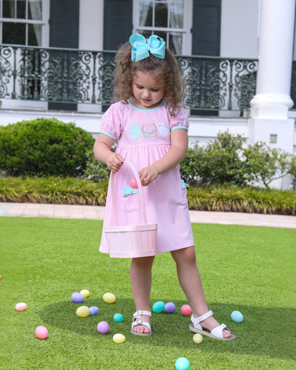 Young girl in a pink dress with colorful eggs on grass in front of a house.